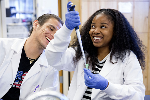 Two people side-by-side in white lab coats. Both are smiling while one is using a 