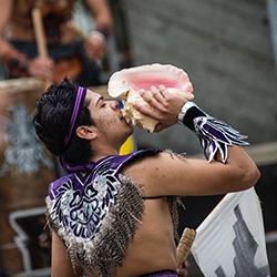 SF State student dressed in purple feathered garment blowing a pink conch
